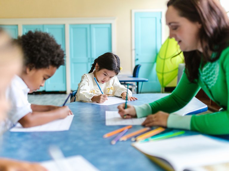 A teacher in a green long-sleeved top sits at a blue classroom table writing on a sheet of paper, with children writing beside her.